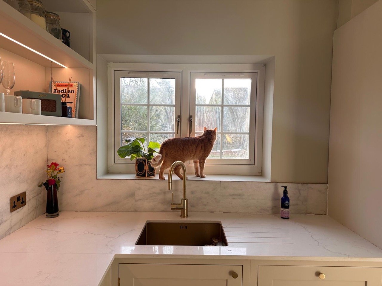 Kitchen worktop and brass sink tap beneath a window, with fitted cabinets and marble-pattern splashback